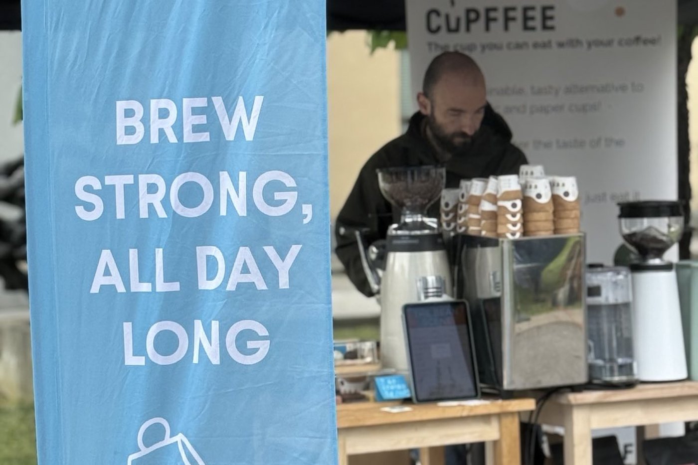 A blue flag reading BREW STRONG, ALL DAY LONG in front of an outdoor coffee stand with grinders and thermal airpots