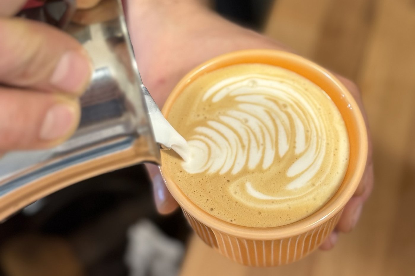 A barista pours milk from a jug into a terracotta cup, shaping latte art